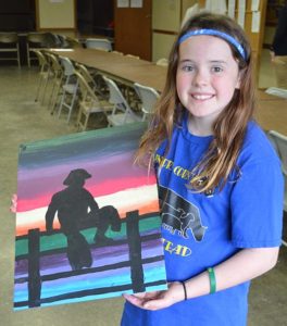 Hope Baysinger shows off her canvas painting of a cowboy sitting on a fence.